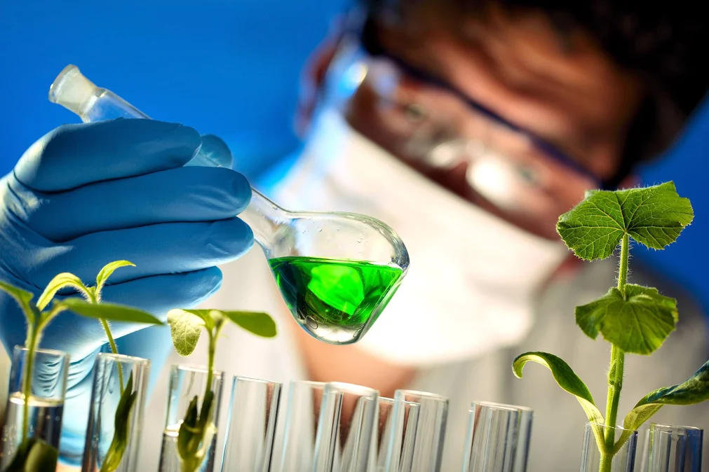 Scientist in protective gear examining green liquid in a flask near plant samples in test tubes, representing biotechnology or agricultural research.